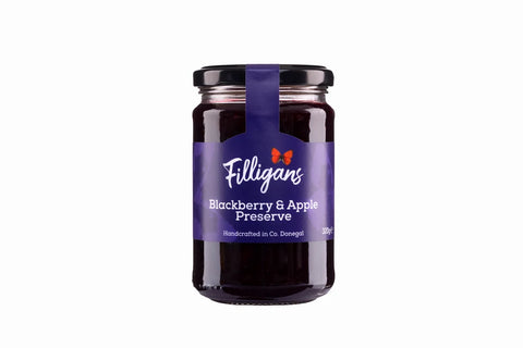 A glass jar of Filligans Blackberry & Apple Preserve on a solid white background. The jar has a dark purple label with white text and a small red bow logo, topped with a black lid and a purple ribbon.