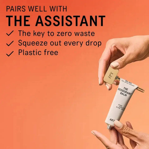A hand holds a white tube of AKT The Deodorant Balm with a gold cap, while another hand unscrews the cap. The background is a solid peach-orange color with promotional text on the left.