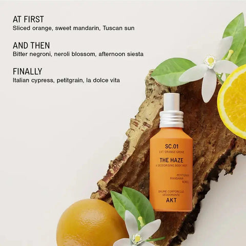 An amber spray bottle labeled 'THE HAZE' by AKT, styled on a rustic wood slice with citrus slices, white flowers, and green leaves against a light neutral background. Text on the left lists scent notes in poetic form.