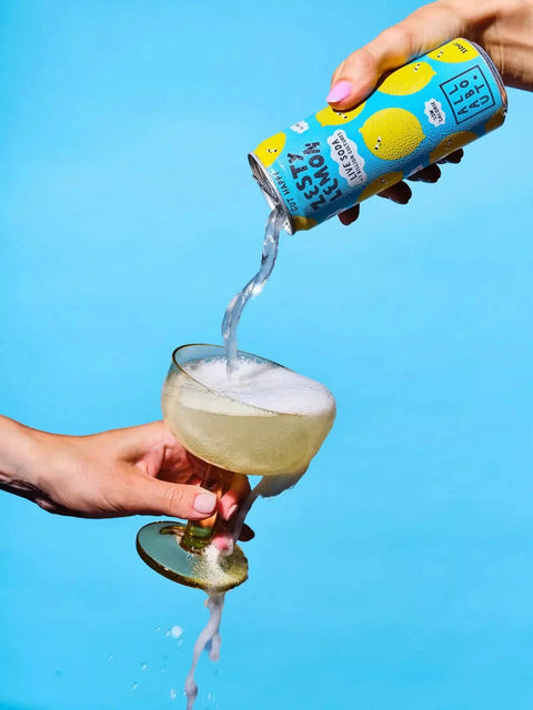 A hand holds a can of Fernet Bruguier Paris Aperitif and pours the clear liquid into a coupe glass filled with a pale yellow drink against a bright blue background.