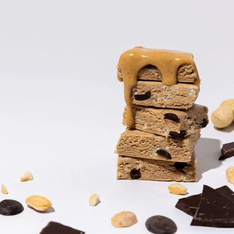 A stack of four chocolate chip cookies topped with a glossy peanut butter drizzle, set on a white background with scattered peanuts, chocolate pieces, and cookie crumbs.