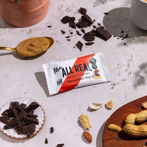 A Mini All Real peanut chocolate chip bar in its packaging, styled on a light marble surface with peanut butter on a spoon, broken chocolate pieces, peanuts, and a small dish of chocolate chunks.