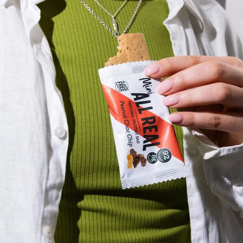 Person holding a 'All Real' peanut chocolate chip bar with a green sweater and white shirt.