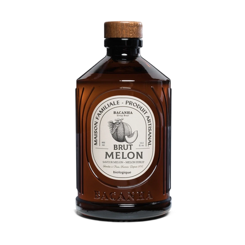 A bottle of Bacanbra Brut Melon liqueur, featuring an amber-brown glass body, a light wood cap, and a cream-colored oval label with black text and a fruit illustration.