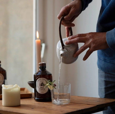 Person pouring liquid from a white ceramic teapot into a glass on a wooden table with candles and bottles in the background.