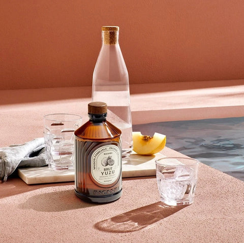 A bottle of Bacardi Blue Yuzu cocktail mix and a clear glass bottle of water are styled on a terracotta-colored surface next to a pool, with ice, a slice of yellow fruit, and folded gray cloth.