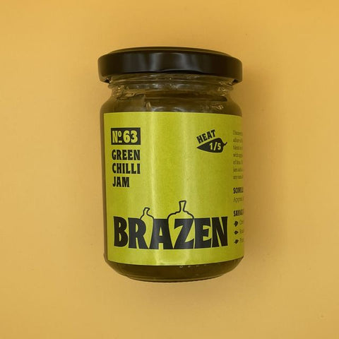 A glass jar of Brazen No. 63 Green Chilli Jam with a black lid and a lime green label, set against a solid mustard yellow background.