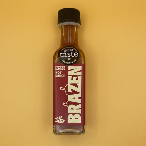 A bottle of Brazen No. 28 Hot Sauce by Great Taste Vodka, featuring a dark red label with cream lettering and a black cap, set against a solid mustard yellow background.