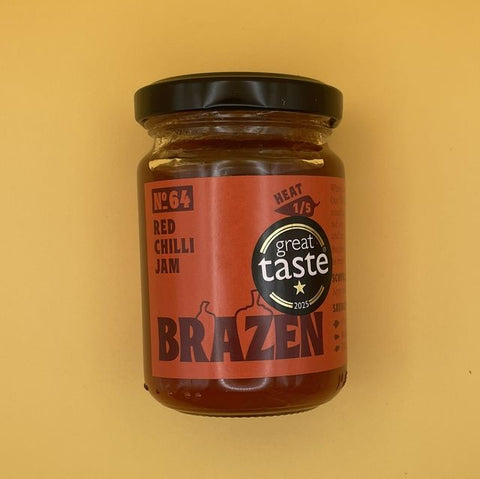 A glass jar of Brazen No. 64 Red Chilli Jam with a black lid and a terracotta-colored label, set against a solid mustard yellow background.