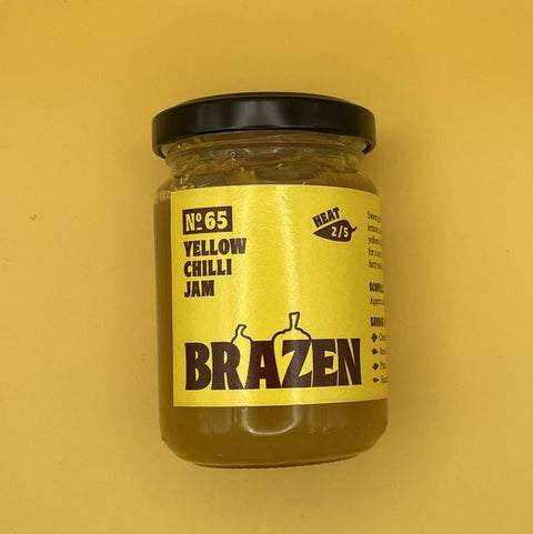 A jar of Brazen No. 65 Yellow Chilli Jam with a black lid and a bright yellow label, set against a solid yellow background.