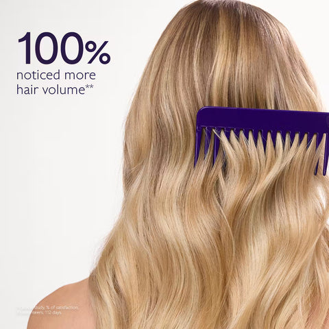 A woman with long, wavy blonde hair seen from the back, using a wide-tooth purple comb. Text on the left reads '100% noticed more hair volume**'.