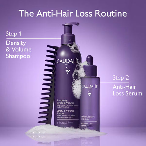 Two purple Caudalie Paris hair care bottles on a lavender background with text reading 'The Anti-Hair Loss Routine' and step labels. The larger bottle is shampoo and the smaller dropper bottle is serum, with a black hair-building brush and foam visible.