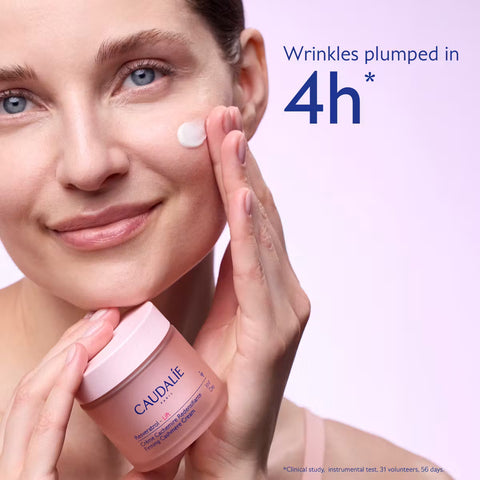 A woman applies cream to her cheek while holding a jar of Caudalie Deep Moisture Cream. The image features a light pink background with promotional text that reads 'Wrinkles plumped in 4h *'.