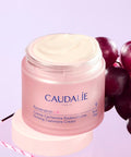 Caudalie Resveratrol-Lift Firming Cashmere Cream in a pink jar with white cream visible on top, styled with dark purple grapes on a light lavender background.