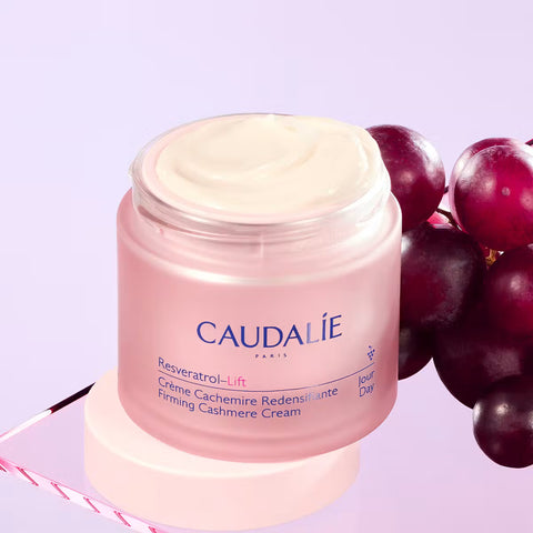 Caudalie Resveratrol-Lift Firming Cashmere Cream in a pink jar with white cream visible on top, styled with dark purple grapes on a light lavender background.