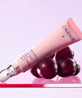 Caudalie Somnatrol-Lis eye gel in a light pink squeeze tube with a metallic silver applicator tip, styled with dark red grape-like spheres on a pink reflective surface against a soft lavender background.