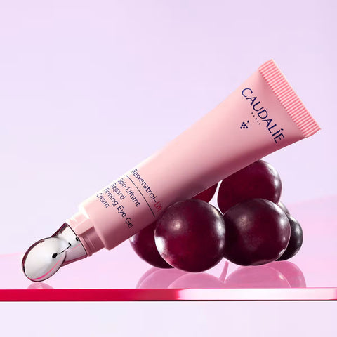 Caudalie Somnatrol-Lis eye gel in a light pink squeeze tube with a metallic silver applicator tip, styled with dark red grape-like spheres on a pink reflective surface against a soft lavender background.