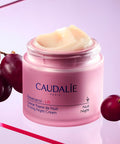 A jar of Caudalie Resveratrol - Lift Firming Night Cream with cream visible at the top, styled with red grapes on a light purple background.