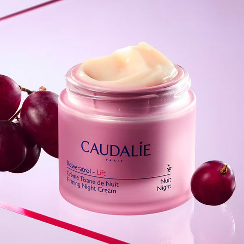 A jar of Caudalie Resveratrol - Lift Firming Night Cream with cream visible at the top, styled with red grapes on a light purple background.
