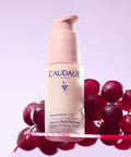 Caudalie Resveratrol Lift Instant Firming Serum in a pale pink pump bottle, styled with red grapes on a pink reflective surface against a lavender background.