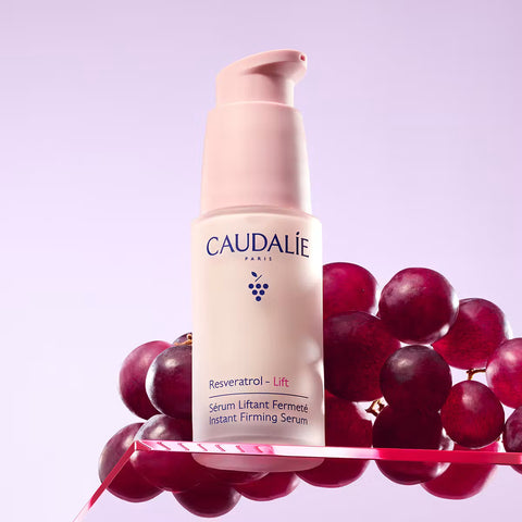 Caudalie Resveratrol Lift Instant Firming Serum in a pale pink pump bottle, styled with red grapes on a pink reflective surface against a lavender background.