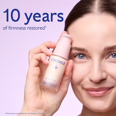 A woman holds a bottle of Caudalie Effacer Lines Visibles 2.5% grapevine serum against a light lavender background, with text reading '10 years of firmness restored'.