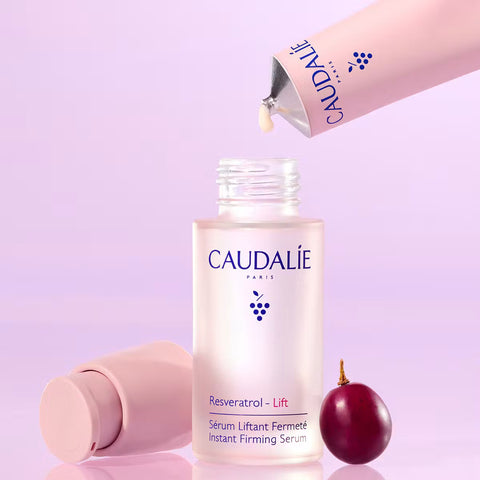 Caudalïe Resveratrol Lift Instant Firming Serum bottle shown on a soft lavender background, with a pink squeeze tube dispensing serum above it and a grape prop at the base.