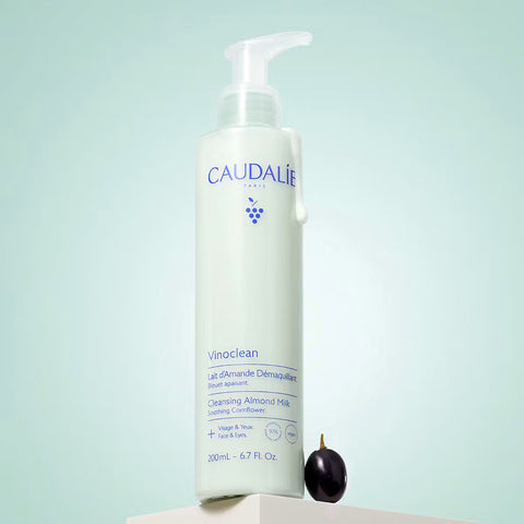 Caudalie Vinoclean Cleansing Almond Milk lotion in a white pump bottle, displayed on a white pedestal next to a dark purple almond with a light mint green background.