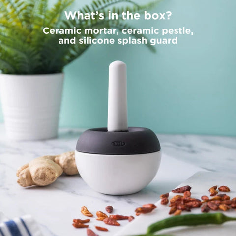 A ceramic food chopper with a white body, dark gray top, and white pestle, shown on a marble surface with ginger, dried red chilies, green chilies, and a potted plant. Text at the top reads 'What's in the box? Ceramic mortar, ceramic pestle, and silicone splash guard.