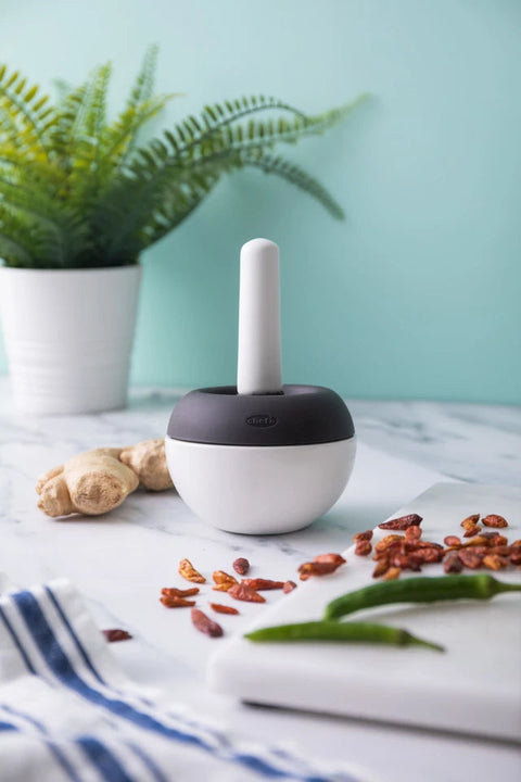 A white and dark gray electric food chopper with a white paddle, set on a marble countertop surrounded by ginger, red chilies, green chilies, and a potted fern against a light blue-green wall.