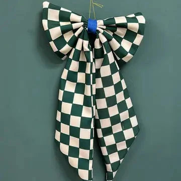 A large fabric bow with a green and off-white checkerboard pattern, tied at the center with a blue ribbon, hanging against a solid dark green background.
