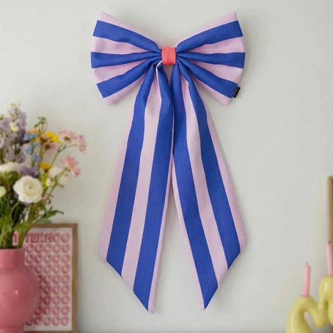 A large fabric bow with vertical blue and light pink stripes, tied with a coral-pink center band, hanging on a plain white wall. A vase of flowers and part of a yellow candle holder are visible at the edges of the frame.