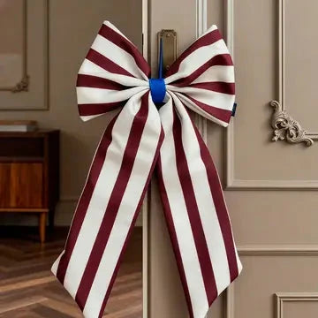 A large decorative bow with vertical burgundy and white stripes, tied with a blue center ribbon, hanging on a beige paneled door in an indoor room.
