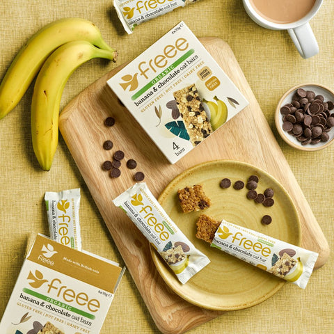 Freee banana & chocolate cut bar snack boxes and wrapped bars arranged on a wooden board with bananas, chocolate chips, a plate with a few bars broken, and a cup of coffee on a yellow textured surface.