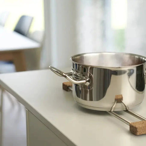 A stainless steel saucepan with a double loop handle resting on a metal trivet with cork pads on a light-colored kitchen countertop.