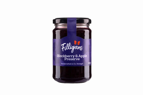 A glass jar of Filligans Blackberry & Apple Preserve on a solid white background. The jar has a dark purple label with white text and a small red bow logo, topped with a black lid and a purple ribbon.