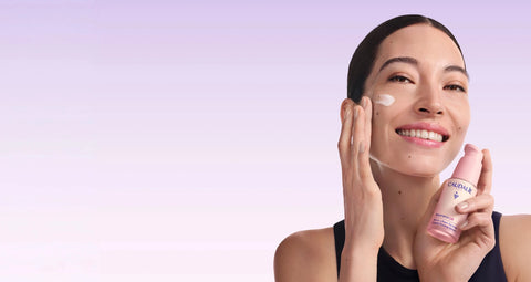 A woman with dark hair pulled back smiles while applying a white cream to her face and holding a pink Vichy Dermo-Cosmetic treatment bottle against a soft lavender gradient background.