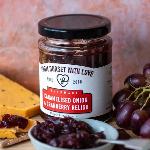 A jar of handmade caramelised onion and cranberry relish from Dorset With Love, styled on a wooden surface with a slice of cheese, a bowl of relish, and grapes.