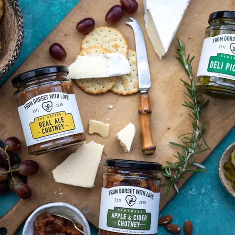 Three gourmet chutney jars from Dorset With Love are arranged on a wooden board with crackers, cheese, grapes, and rosemary on a turquoise surface. The jars include Real Ale Chutney, Apple & Cider Chutney, and Deli Pickles.