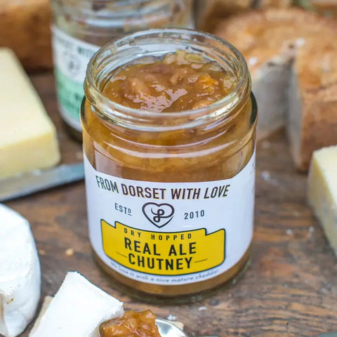 A glass jar of 'From Dorset With Love' Real Ale Chutney on a rustic wooden board, surrounded by cheese wedges and sliced bread.