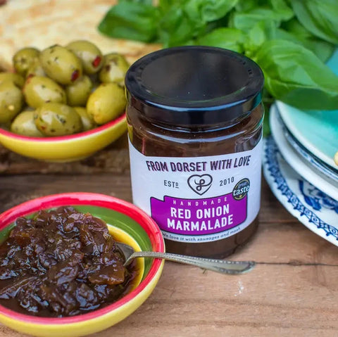 A jar of handmade red onion marmalade from Dorset with Love sits on a wooden table, surrounded by a bowl of chopped red onion, green olives, and fresh basil leaves.