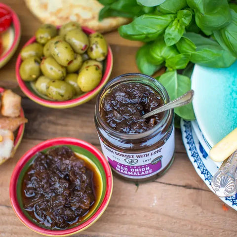 A glass jar of 'Her Onion' marmalade by Sweet Biscuit with Love sits on a rustic wooden table surrounded by fresh ingredients, including green olives, basil, and bread.
