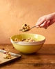 A person's hand holding a spoonful of cereal over a yellow bowl on a wooden table, with a slice of toast nearby.