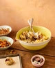 A yellow bowl filled with cereal and topped with syrup, set on a wooden table with small side dishes and a spoon in a styled breakfast scene.
