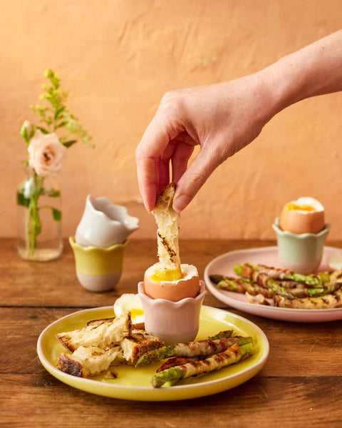 A hand is dipping a piece of toast into a soft-boiled egg on a small pink ceramic egg cup, set on a yellow plate with other breakfast items on a wooden table. In the background, another matching pink egg cup holds a hard-boiled egg on a plate with asparagus and bacon.