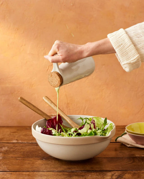 Person pouring salad dressing from a ceramic container onto a salad in a white bowl on a wooden table.
