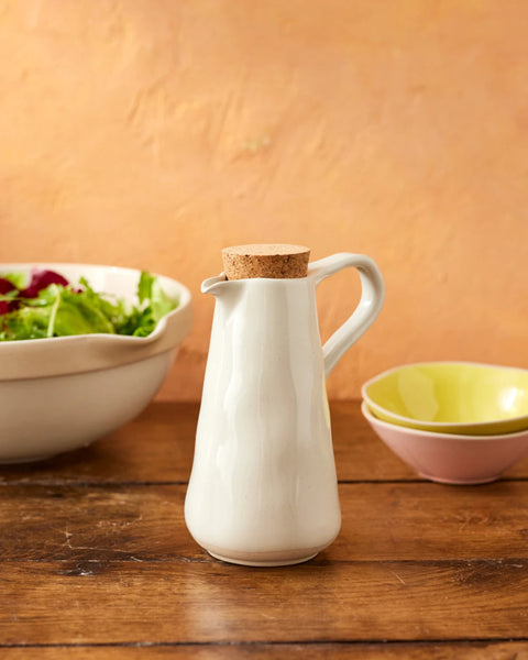 White ceramic pitcher with cork lid on a wooden surface with bowls of salad and fruit in the background.