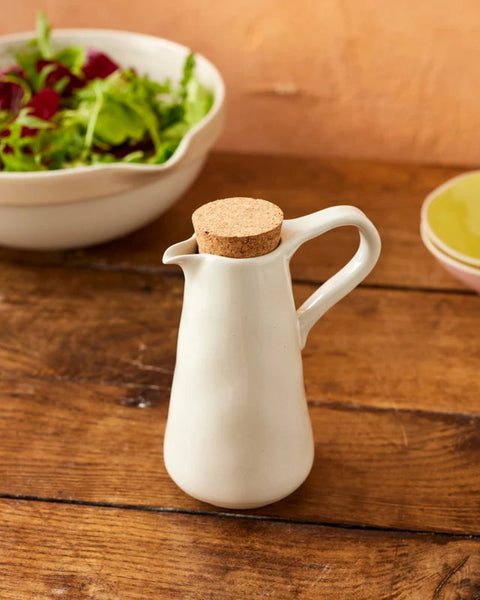 A small white ceramic pitcher with a cork stopper, set on a rustic wooden table with a bowl of salad in the background.