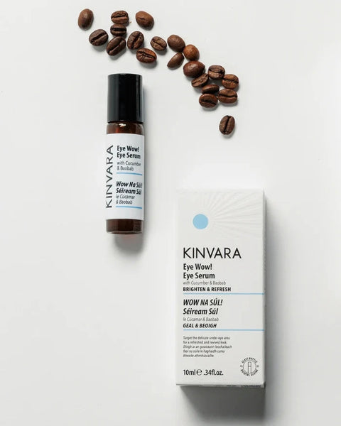 Kinvara Wow Wow! Eye Serum shown with its white and blue retail box and a small amber roller bottle, arranged on a white background with scattered coffee beans.