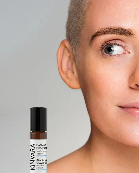 A close-up of a woman's face with a KINVARA Eye Woe! Eye Serum bottle placed near the lower left on a light gray background.